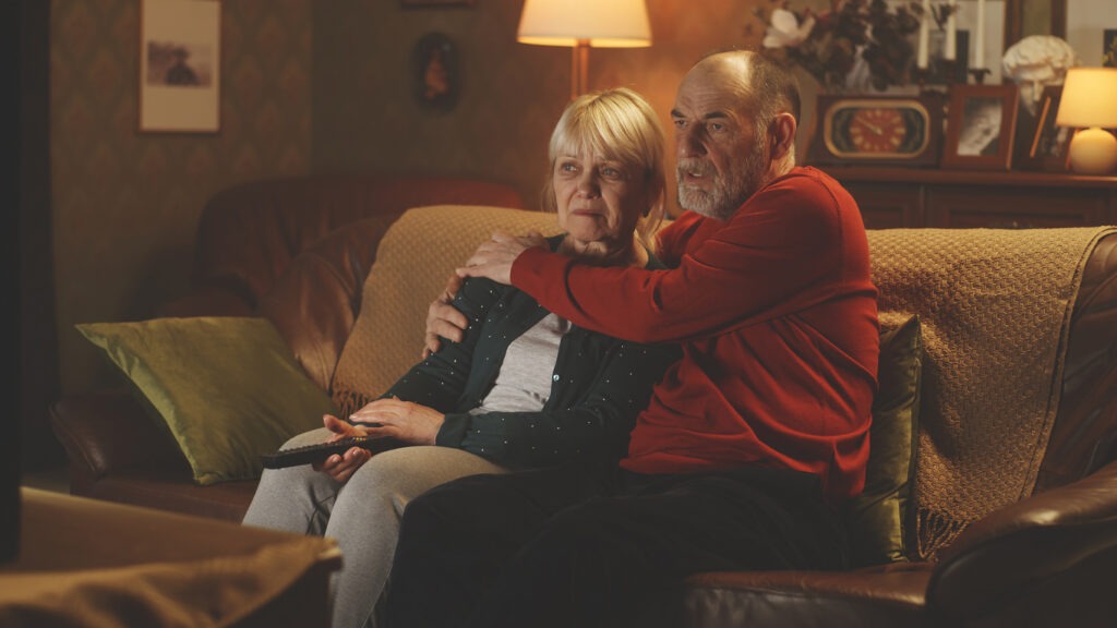 Scared senior husband clasping hands while sitting on the couch and watching a film in the obscure living room at night at home