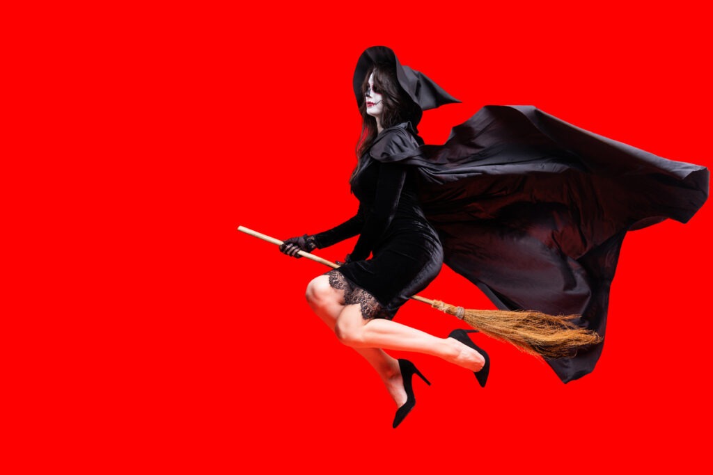 Woman in Halloween costume wears black witch hat, long cloak with creative sugar skull makeup. On a red isolated background fly on a magic broom. Concept Los Muertos poster party or La Calavera Catrina