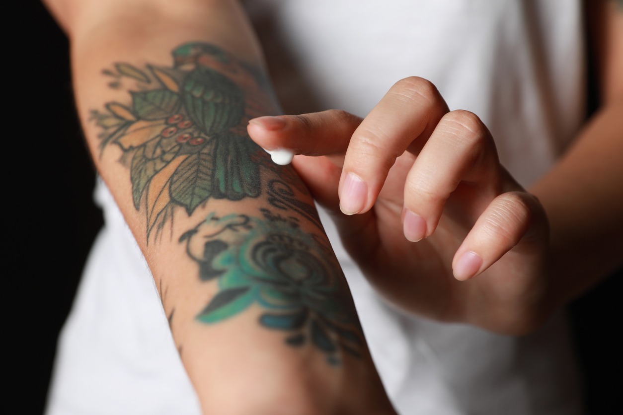 Woman applying cream on her arm with tattoos against black background, closeup