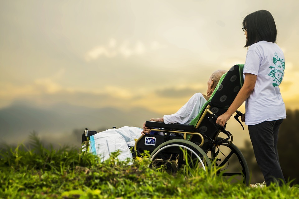 An aged woman resting on a wheelchair overlooking a gorgeous vista with a caretaker pushing the wheelchair