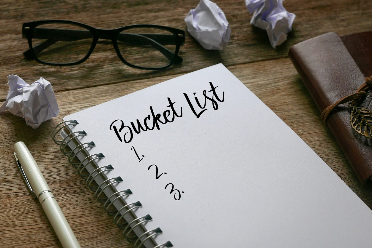 Pen, trash paper, sunglasses, and notebook written with Bucket List on the wooden background