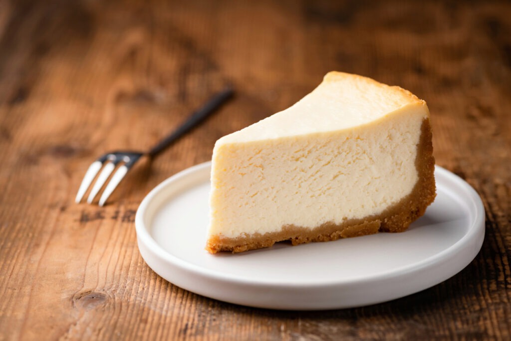 Cheesecake slice, New York style classical cheesecake on wooden background. Slice of tasty cake on a white plate served with dessert fork