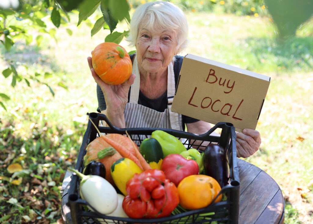 An elderly gray-haired woman sells fresh seasonal vegetables at a local farmers' market. Local business support. Buy local non-GMO agricultural products