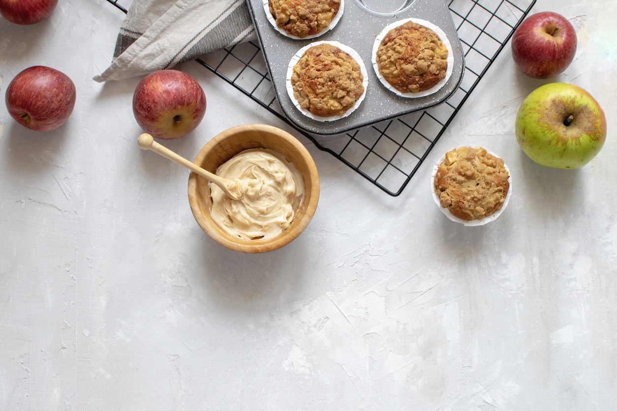 Homemade baking - flat lay of freshly baked apple muffins on a cooling rack, organic apples, spiced butter in a wooden bowl, white background, top view