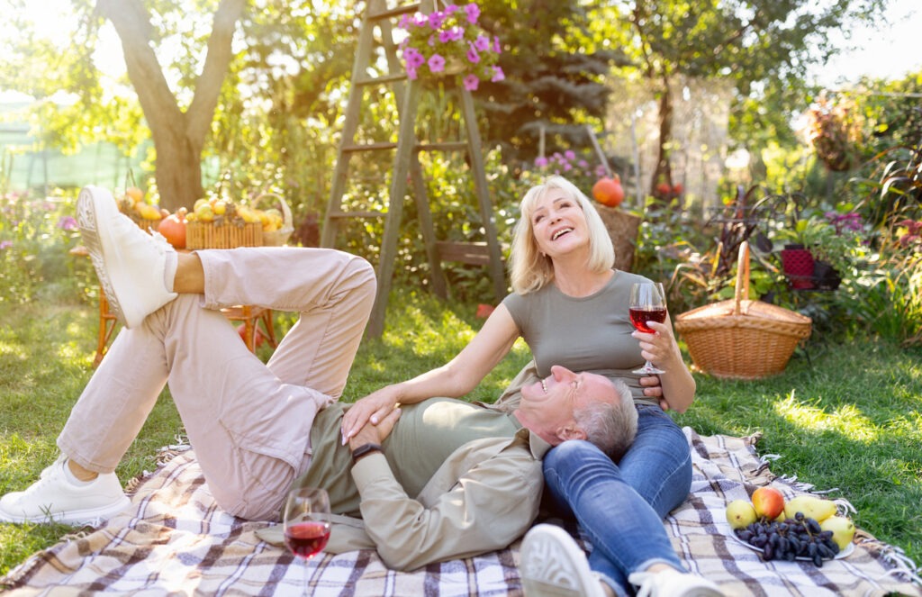 Romantic senior spouses enjoying a picnic together, resting on a blanket in the garden and drinking red wine, chatting and laughing, enjoying the weekend outdoors, free space