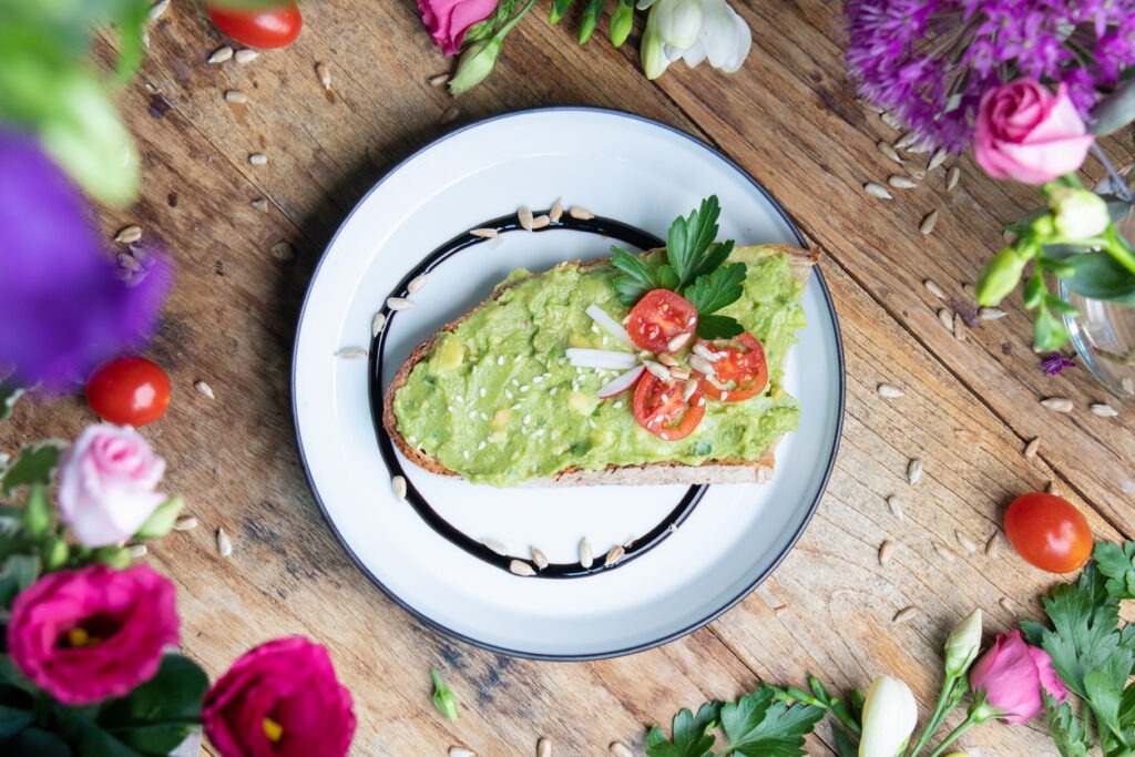 A top view of an avocado toast with tomatoes and seeds on a wooden table among flowers