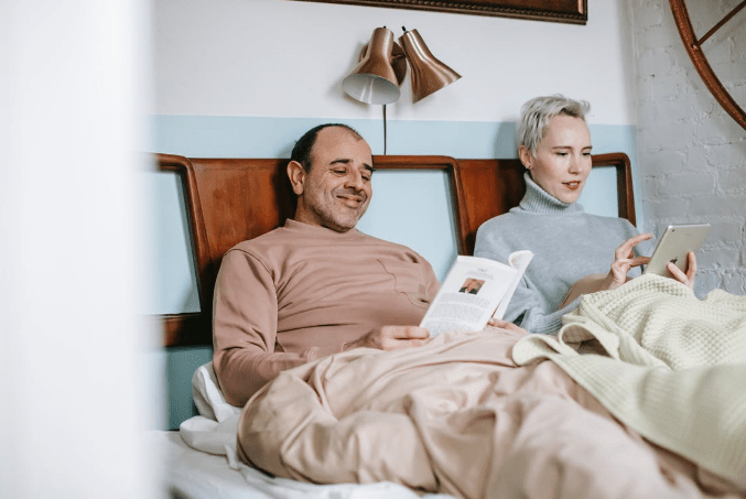 adult-diverse-married-couple-reading-book-and-using-table-in-bed