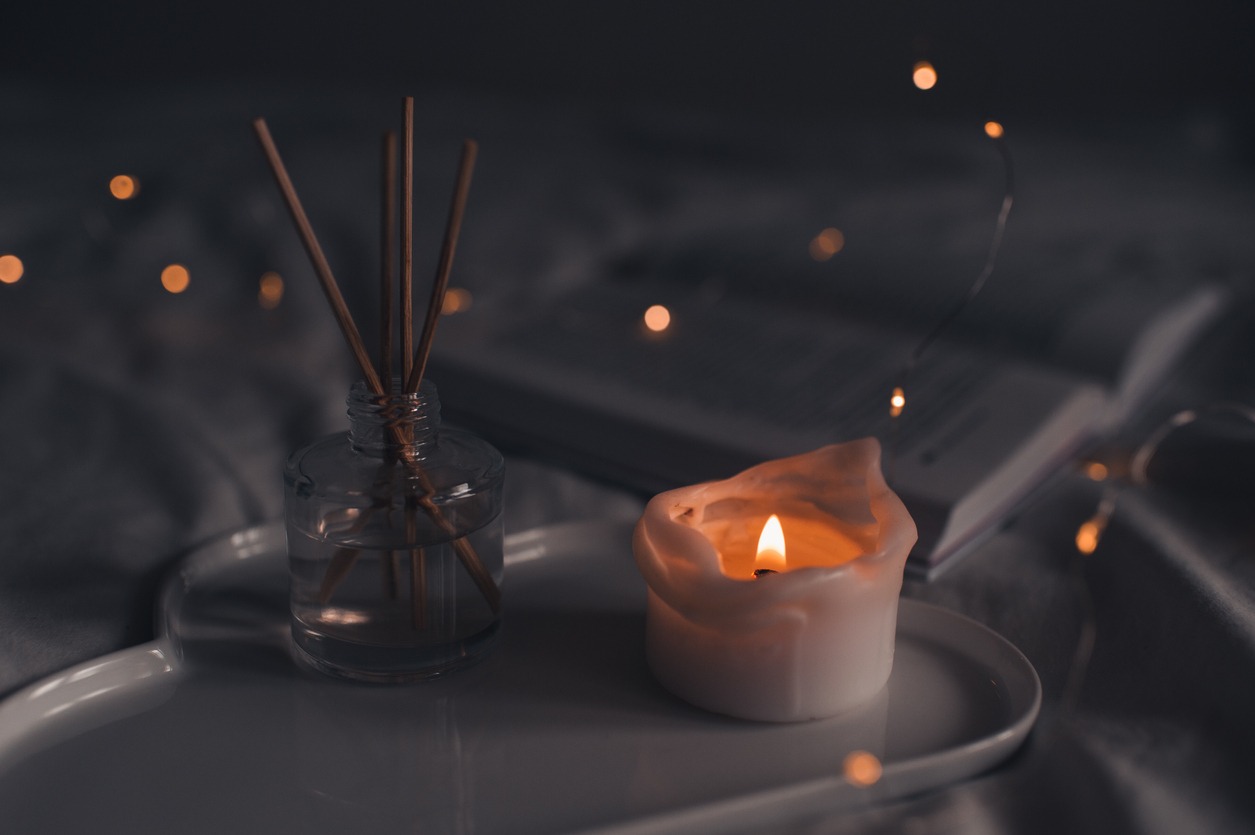 A burning scented candle besides bamboo sticks in a glass bottle on a tray in a bed