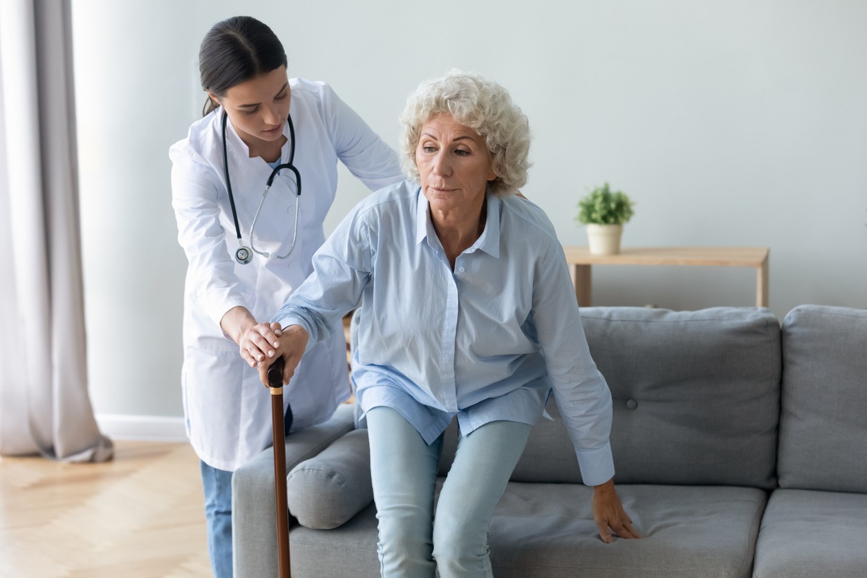 caregiver helping a woman get up from the sofa while holding a walking stick